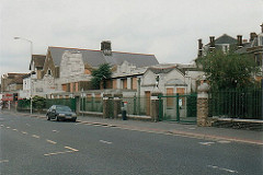 A light grey stone building behind green railings, viewed from the other side of a wide but quiet road.  All of the windows are boarded up with what looks like chipboard.