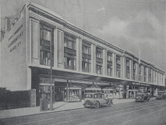 A black-and-white image of a long three-storey building with a columnar look on the upper two floors and a suggestion of individual glazed shopfronts on the ground floor.