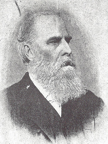 A black-and-white photograph of a white man with a receding hairline, a prominent browline, and a full white beard and moustache.  He is wearing a white shirt and dark jacket, and looking somewhat stern.