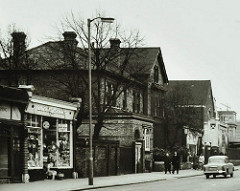 A black-and-white photograph showing a 2–3-storey brick house with peaked roofs and protruding extensions.  A few people are on the pavement outside, and an old-fashioned car is in the road.  A small terrace of shops begins to the left.