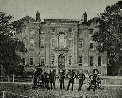 A black-and-white photograph of a three-storey building with a grand arched entrance.  Trees are to either side.  Nine people are posing in front of the building, all fairly tall and thin, and all dressed in uniform; several of them are leaning on the small cannons drawn up on each side of the groups.