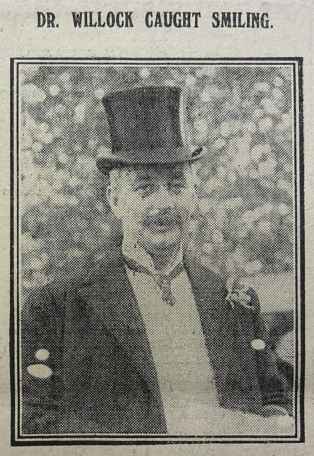 A black-and-white photo of a white man with a moustache, wearing a top hat and a dark jacket with what looks like a flower in a buttonhole.  He is looking towards the camera and possibly smiling (the caption reads: “Dr. Willock caught smiling.”)