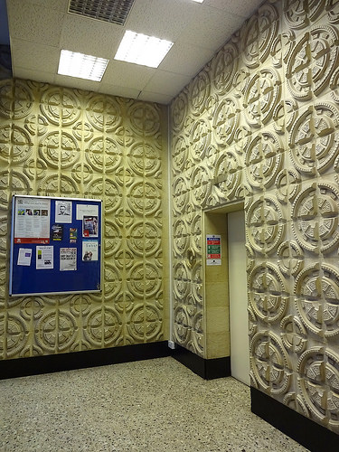 The corner of a hallway, with a lift and noticeboard. The walls are covered with ornate quartered-circle mouldings.