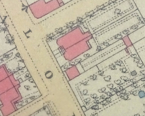 Extract from a hand-coloured map showing houses (shaded in pink) with gardens including markings indicating trees and paths, on either side of a main road with two side roads.