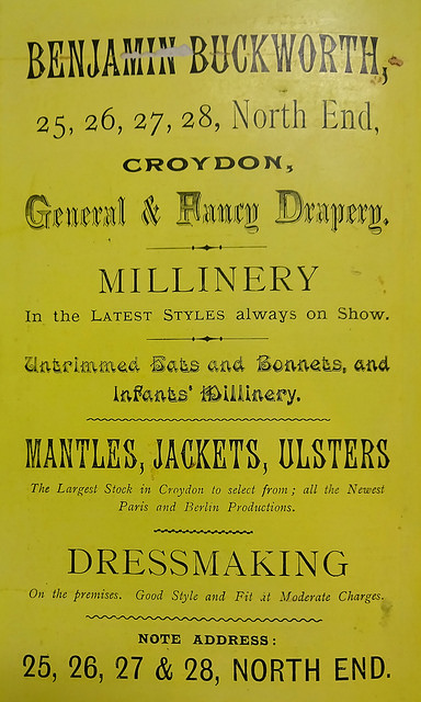 An advert in a variety of fonts, printed in black on shiny yellow paper, headed “Benjamin Buckworth, 25, 26, 27, 28, North End, Croydon, General & Fancy Drapery.”  Also advertised are millinery, untrimmed hats and bonnets, mantles, jackets, and ulsters, as well as “Dressmaking On the premises”.