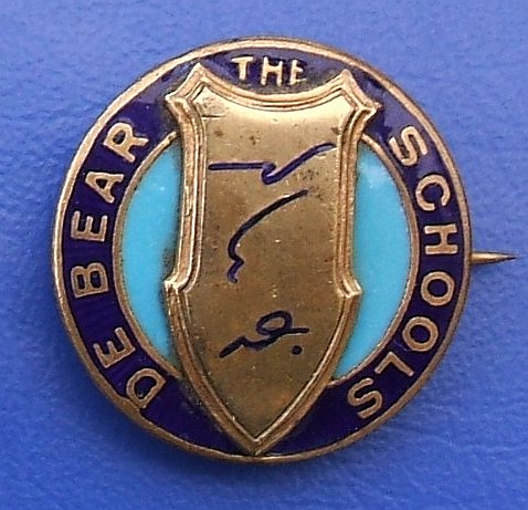 A round metal/enamel badge with the end of its pin just visible sticking out at the right-hand side.  An elongated bronze shield is in the middle, with three shorthand symbols on it, and “The De Bear Schools” is printed in capitals around the edge.