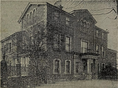 A black-and-white photo of a three-storey house with pillars at the entrance and a wall running away from it on one side.  The photo is taken from a newspaper so is somewhat grainy.