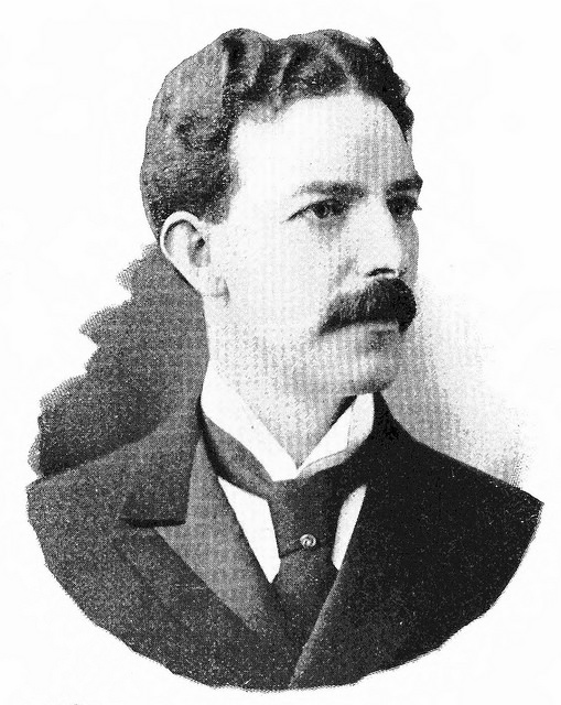 Black-and-white photo of a youngish white man with short, dark, wavy hair and a substantial moustache, looking off towards the right of the image.  He is wearing a white shirt, dark suit jacket, and dark tie.