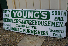 A long enamelled sign resting on the ground against a wall.  The text is green on a white background, and reads: “Young’s / North End.  South End.  Removers & Warehousemen.  Complete House Furnishers.  Estabd 1864.  Estimates Free.”