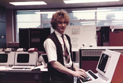 A white person in masculine clothing (checked tie, short-sleeved white collared shirt, brown waistcoat) looking towards the camera in a room full of computer terminals.  Their hands are on the keyboard of one of the terminals.  Daylight and the chimneys of another building are visible through a window behind.