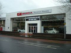 A large,
white, blocky building with large plate-glass windows showing cars
parked inside.  A sign above the windows reads “HONDA / Newton
Honda”.