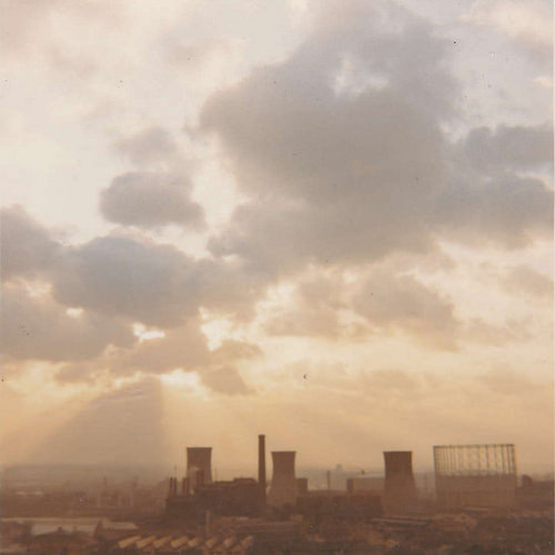 A hazy photo showing the sun behind clouds with power station cooling towers and a gasholder below.
