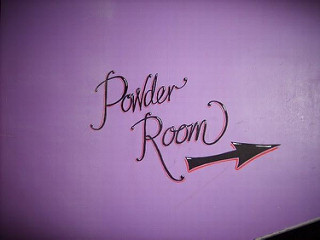 A pinkish-purple wall with the words “Powder Room” painted on in black cursive.  Below is an arrow pointing slightly upwards.