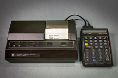 Two pieces of 1980s-looking hardware with black plastic cases, connected together with thin black cables.  The one on the left is larger, and has a narrow piece of paper coming out of it with “Hewlett Packard / 821621A / Thermal Printer” printed on it in all-caps, and the one on the left has a blank one-row display above several rows of buttons labelled with letters, numbers, and abbreviations of mathematical functions such as sine, cosine, and tangent.