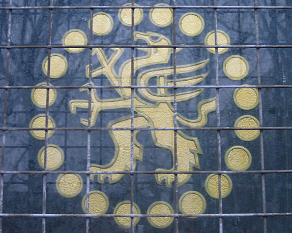 A circular logo embossed on a window behind a thin grille.  In the centre is a stylised griffin, surrounded by a circle of smaller circles (representing coins).