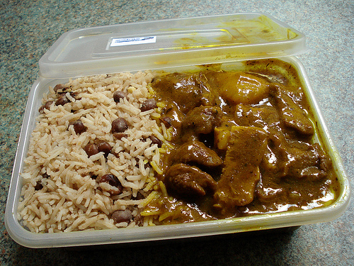 A rectangular plastic takeaway tub with rice & peas in the left side and curry goat in the right.