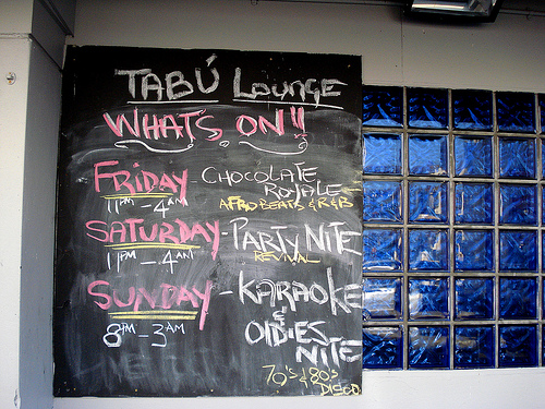 A blackboard with chalked writing in red, yellow, and white: “Tabú Lounge / Whats On!! / Friday 11pm–4am Chocolate Royale Afrobeats & R&B / Saturday 11pm–4am Party Nite Revival / Sunday 8pm–3am Karaoke & Oldies Nite 70’s & 80’s Disco”.  The dark blue tiling mentioned earlier is visible to the side.