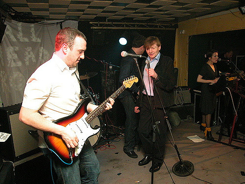 Four white people on stage in a low-roofed venue.  One is playing a guitar, one is holding a microphone, one is playing a keyboard, and one is turned away from the camera and partly obscured by the microphone-holder.