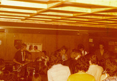 A yellow-tinged photo of five young white people playing on stage in a low-ceilinged room with an audience crowded up together in front of the stage.