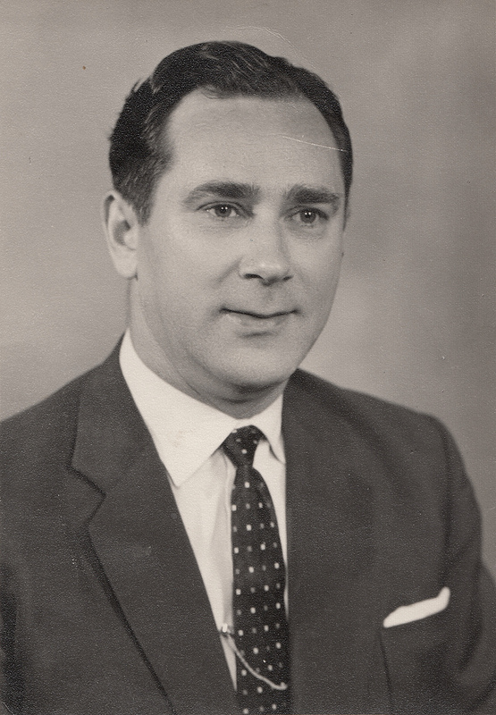 Monochrome photo of a white man with neat dark hair and a wistful look, wearing a dark suit with white pocket square, white shirt, and dark tie patterned with small white dots.