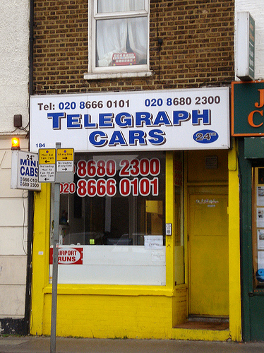 A small terraced shopfront painted in mustard yellow, with a large white sign above reading “Telegraph Cars”.  Two phone numbers are shown prominently in the front window.  There is a recessed door to the right, again painted mustard yellow, and an intercom on the front of the shop next to it.