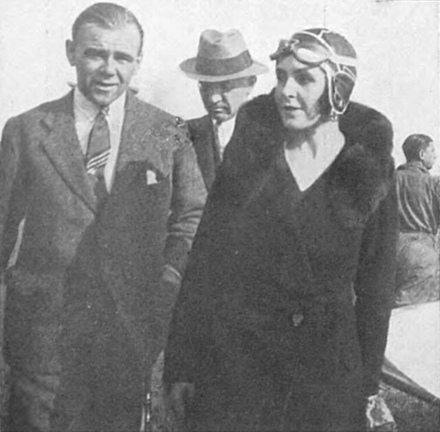 A black-and-white photo of two people (with another behind them, mostly obscured) standing or walking towards the camera.  The one on the left is wearing a suit with a tie and buttoned-up jacked, and the one on the right is wearing a fur coat and aviator’s cap and goggles.