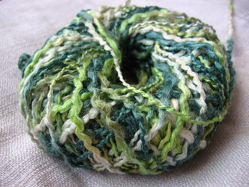 A small, round, flat ball of bouclé-style yarn in variegated shades of cream and green, lying on a woven white cloth.
