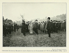 A side-on view of men in bowler hats and dark coats standing in long lines in a field.  A short man in a military hat and with a long pale double-breasted coat is walking between one of the lines towards the camera, followed by at least two more bowler-hatted men.
