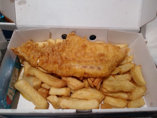 A portion of battered fish, sitting on top of a layer of thick-cut chips in a cardboard box.  A sachet of tartare sauce is tucked to one side.