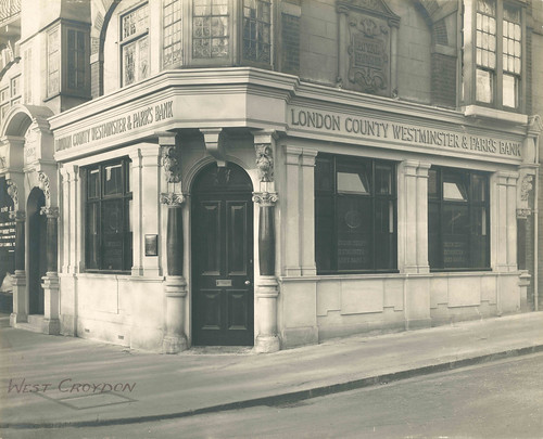 A black-and-white photo of a ground-floor end-of terrace property with an arched doorway at the corner and large rectangular windows along both sides.  An embossed sign running above the windows reads “London County Westminster & Parr’s Bank”.  “West Croydon” has been handwritten in the lower left corner of the photo.