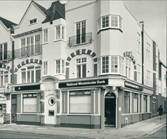 Two shop premises at the end of a terrace, with uniform “National Westminster Bank” branding across both.  The entrance is at the corner of the terrace, with the number “222” above.  There is arched decorative brickwork on the first floor, and wrought-iron balconettes on the second.