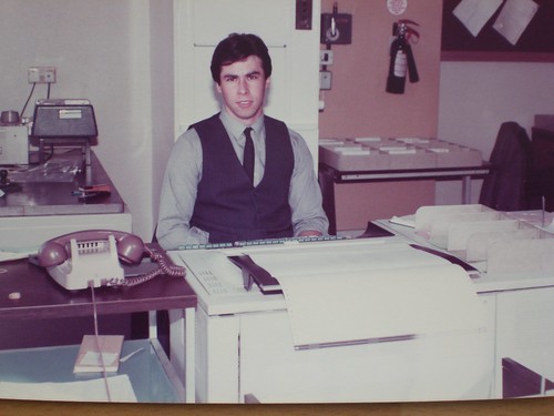 A young white man wearing a light-blue shirt with dark tie and waistcoat, sitting behind some sort of computer-style machine with a keyboard and printout slot.  An old-fashioned telephone with handset and spiral cord is on a desk to one side, and index-card boxes are visible in the background.
