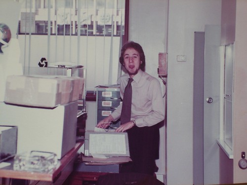 A different young white man wearing a light-brown shirt with a dark patterned tie.  He is standing at the side of a desk with some files and folders on top of it, and poking his tongue out at the camera.  A card index cabinet is behind him, and beyond that is a window with open blinds, through which the mouldings of the Zodiac Court frontage are just visible.