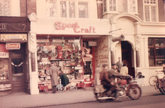 The same shop again, but now the photo is in colour, albeit with a very reddish cast.  The “SportCraft” sign is still present, as is the painted sign for L F Godfrey’s coals at the side of the entrance.  The shop window is covered with toys and games, apparently hanging on hooks or mounted on shelves, to the point where it seems unlikely anyone would be able to see inside.  An adult and a child are looking at the window display, a couple of other adults are standing on the pavement, and two people are whizzing past on a scooter.