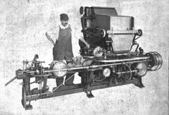 A black-and-white photo of a person with an impressive moustache, wearing a cap and dungarees and standing with one hand on their hip behind a long machine which seems to involve quite a lot of rollers.