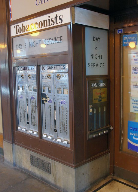 Three old-fashioned vending machines inlaid into two sides of a protruding wall.  A sign at the top reads “Tobacconists”, and others just below this read “Day & Night Service” and “Cigarettes”.  The machines themselves are plain metal, flush with the wall, and have coin slots at the tops.  Partially-removed stickers and plaques advertise John Player Special and Benson & Hedges cigarettes.