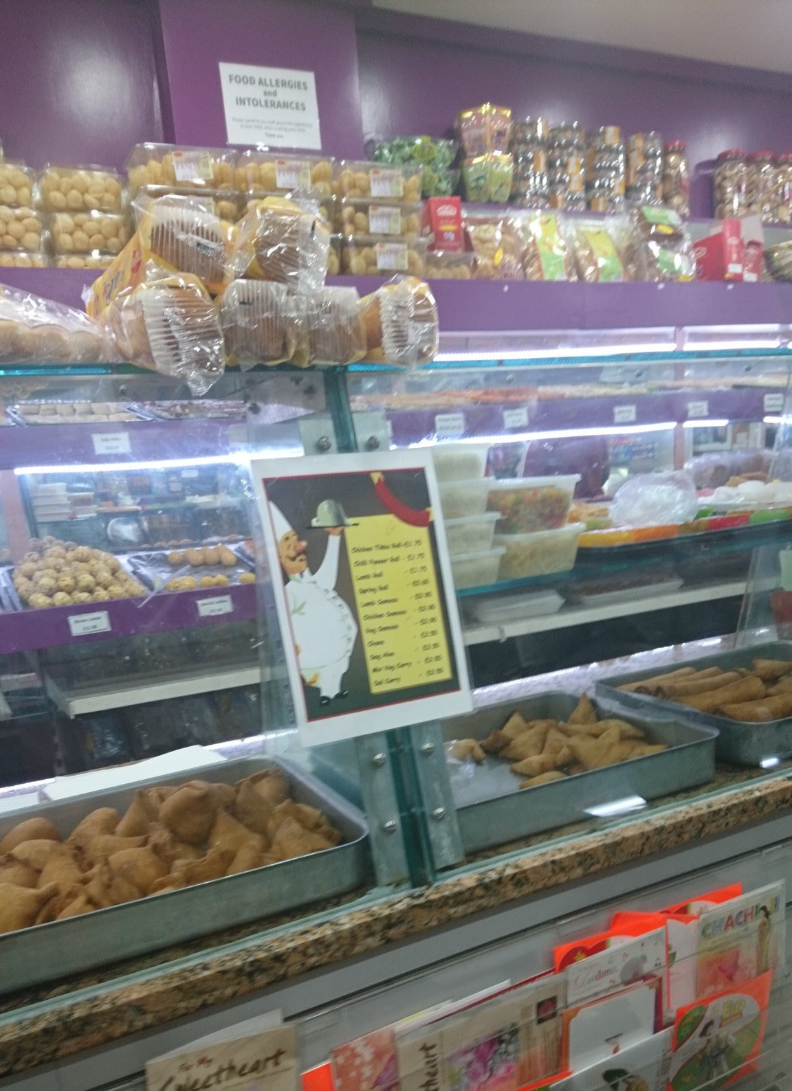 A glass-fronted display counter with trays of samosas.  Behind are shelves lined with Indian sweets.