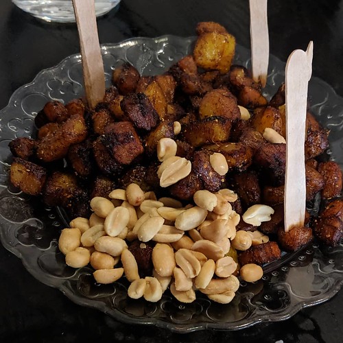 A clear patterned plate piled with dark brown cubes of plantain and a handful of roasted peanuts.  Small wooden forks are stuck upright in the pile.