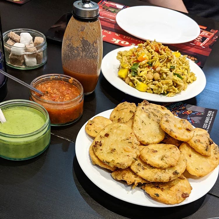 A plate of thin oval fritters in the foreground, with another plate of mixed puffed rice, chickpeas, and cubed potatoes behind.  Small glass dishes on the side hold red and green sauces.