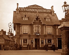 A monochrome photo of the same pub; only the original side is visible, but there is clearly an extension to the right.  A sign on the roof reads “The Fox & Hounds.  City of London Brewery Co”, while lower down are other signs reading “Foreign Wines” and “J. L. Denton”.
