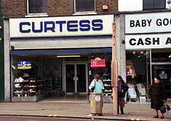One-and-a-half terraced shopfronts. The full shopfront is on the left, with a sign above reading “Curtess” and a recessed entrance with two glass-fronted displays of shoes on either side. Three people are standing on the pavement in front of the shops.