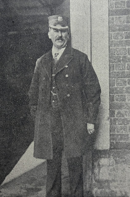 A black-and-white photo of a white man with a moustache, wearing a dark uniform and peaked cap, and standing against a brick wall.