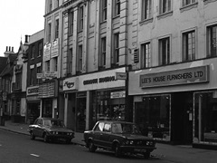 A black-and-white photo of an oblique view onto a row of shops of varying architectural styles. The bulk of the picture is taken up by a tall building in the foreground. The closest shop has a sign reading “Lee’s House Furnishers Ltd”, and the one next to it has one reading “Argos Catalogue Showroom”.