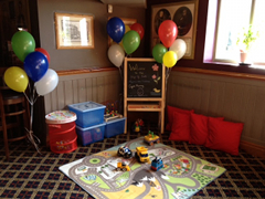 A corner inside a pub, with a diamond-patterned carpet on the floor overlaid with a town-themed playmat. Toy vehicles are placed on the playmat, and plastic boxes presumably containing other toys are against the walls. A blackboard reads: “Welcome to the Ship of Fools [text too small to read]”. Coloured balloons are attached to the blackboard and a nearby stool.