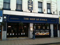 A shop-conversion pub in the middle of a terrace. A sign above reads “Wetherspoon / The Ship of Fools / Free House”. Posters in the windows advertise meals costing around £6. A person is standing in the street looking at the menu posted next to the door.