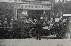 A street scene with old-fashioned vehicles in the foreground, several people milling around, and a shop in the background with a sign reading “Books Purchased.  W. J. Radlett.”