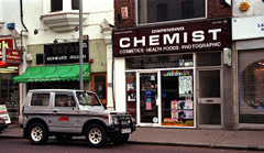 A terraced shopfront with a sign above reading “Dispensing Chemist” in white text on a dark brown background. Posters and decals in the window advertise things like spectacles, nappies, ear piercing, and hair products. A grey SUV is parked outside. To the left of the shop is a small restaurant with a green canopy and a sign above reading “Papa’s Ristorante Italiano”.