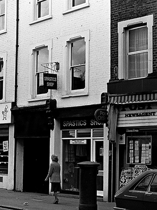 A section of a shop terrace, with an alleyway to the left, a narrow shopfront with “Spastics Shop” above it in the middle, and a post office to the right. A postbox stands outside the post office, and a pedestrian is walking past.