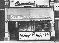 Black-and-white photo of a ground-floor shop with a sign above reading “The Cavendish for Refreshments”.  The canopy over the window reads “Senior Service Satisfy”, and a large riser below has the phrase “Player’s Please” in cursive.