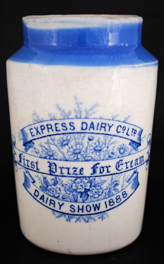 A white and blue ceramic pot against a black background. Writing on the pot reads: “Express Dairy Co Ltd / First Prize for Cream / Dairy Show 1888”. The writing is on stylised scrolls against a background of flowers.